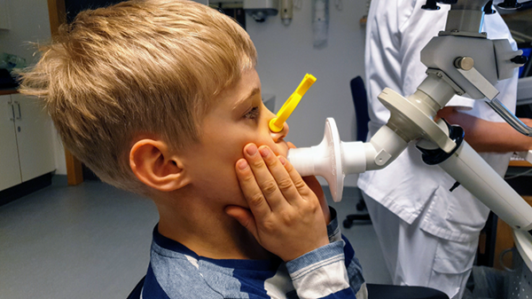 ett barn med en klämma för näsan blåser i ett rör kopplat till en maskin, vårdpersonal i bakgrunden. foto. 