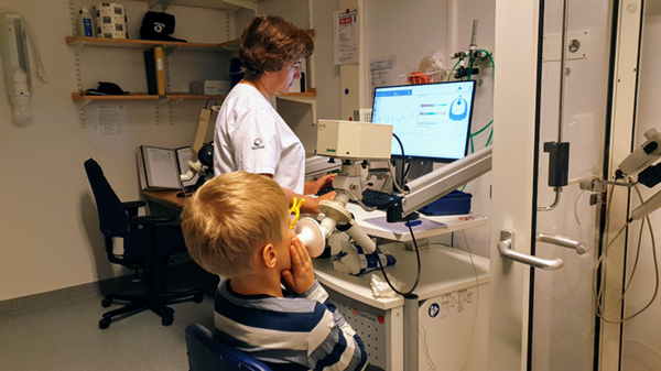 vårdpersonal står vid en dator medan ett barn blåser i ett rör kopplat till en maskin. foto. 
