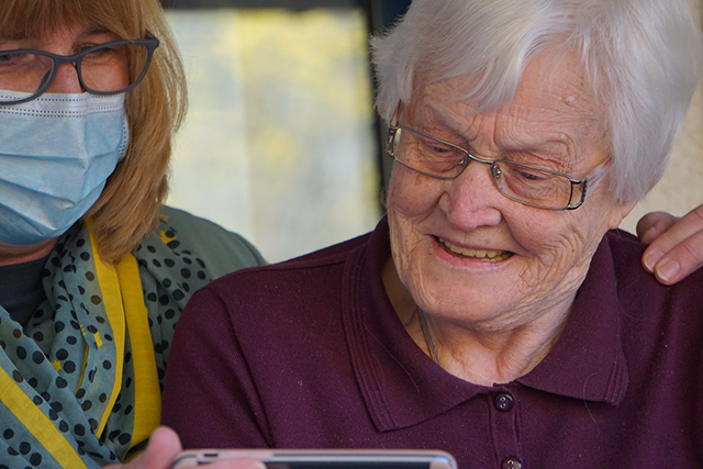 person med ansiktsmask visar en äldre kvinna något på en mobiltelefon. Foto. 